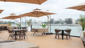 Un espace de restauration en plein air, avec des tables et des chaises placées sous des parasols, offrant une vue sur le bord de l’eau et les bâtiments qui se dressent à l’arrière-plan.