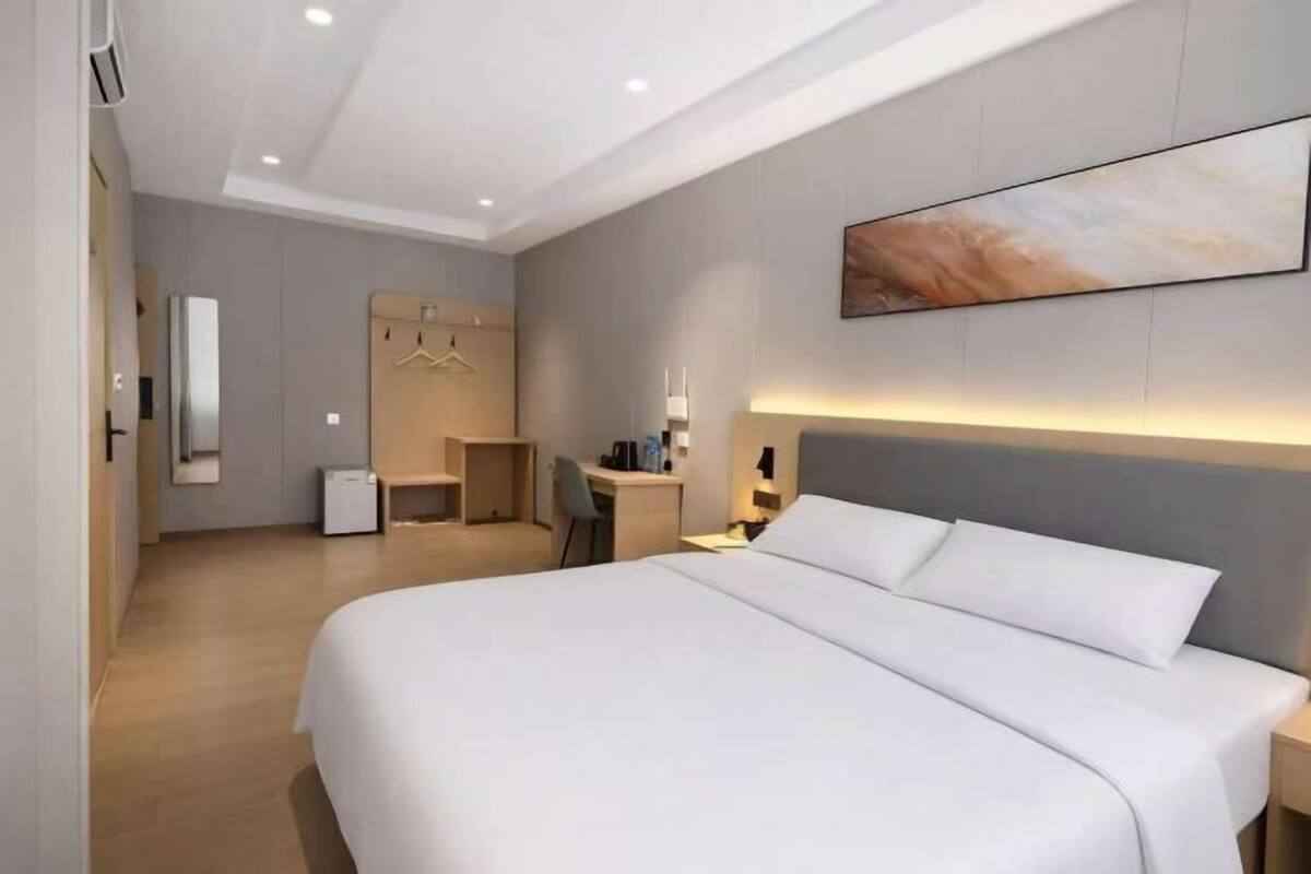 A modern hotel room with a large bed, a desk, a chair, a wardrobe, and a painting on the wall.