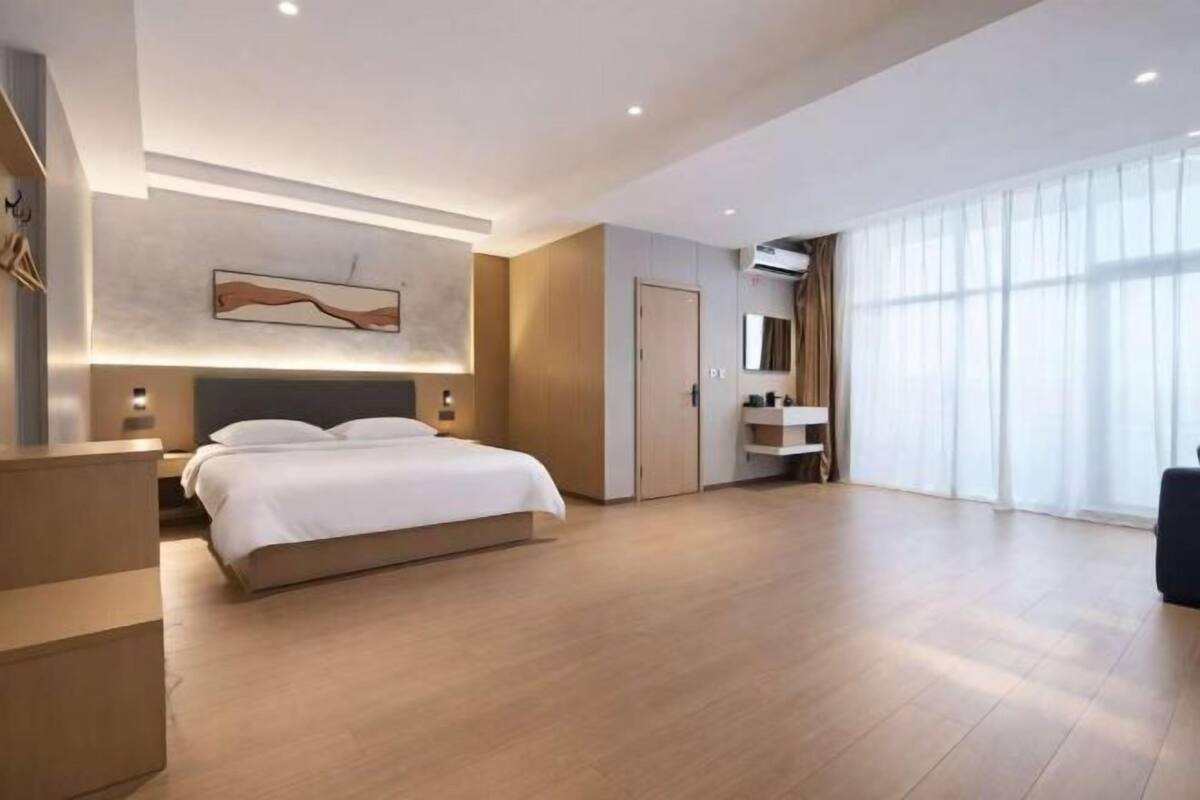 A modern hotel room with a large bed, a bedside table, a wall-mounted artwork, a built-in wardrobe, and a large window with curtains.