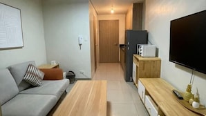A modern living room with a grey sofa, a wooden coffee table, a flat-screen TV mounted on the wall, and a kitchen area in the background. - Coast Apartment (Pasay)