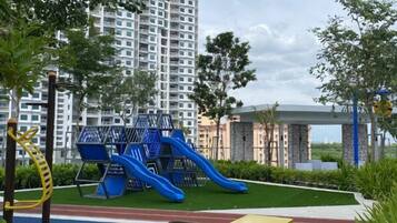 A playground with slides and climbing structures, surrounded by greenery and a tall residential building in the background.