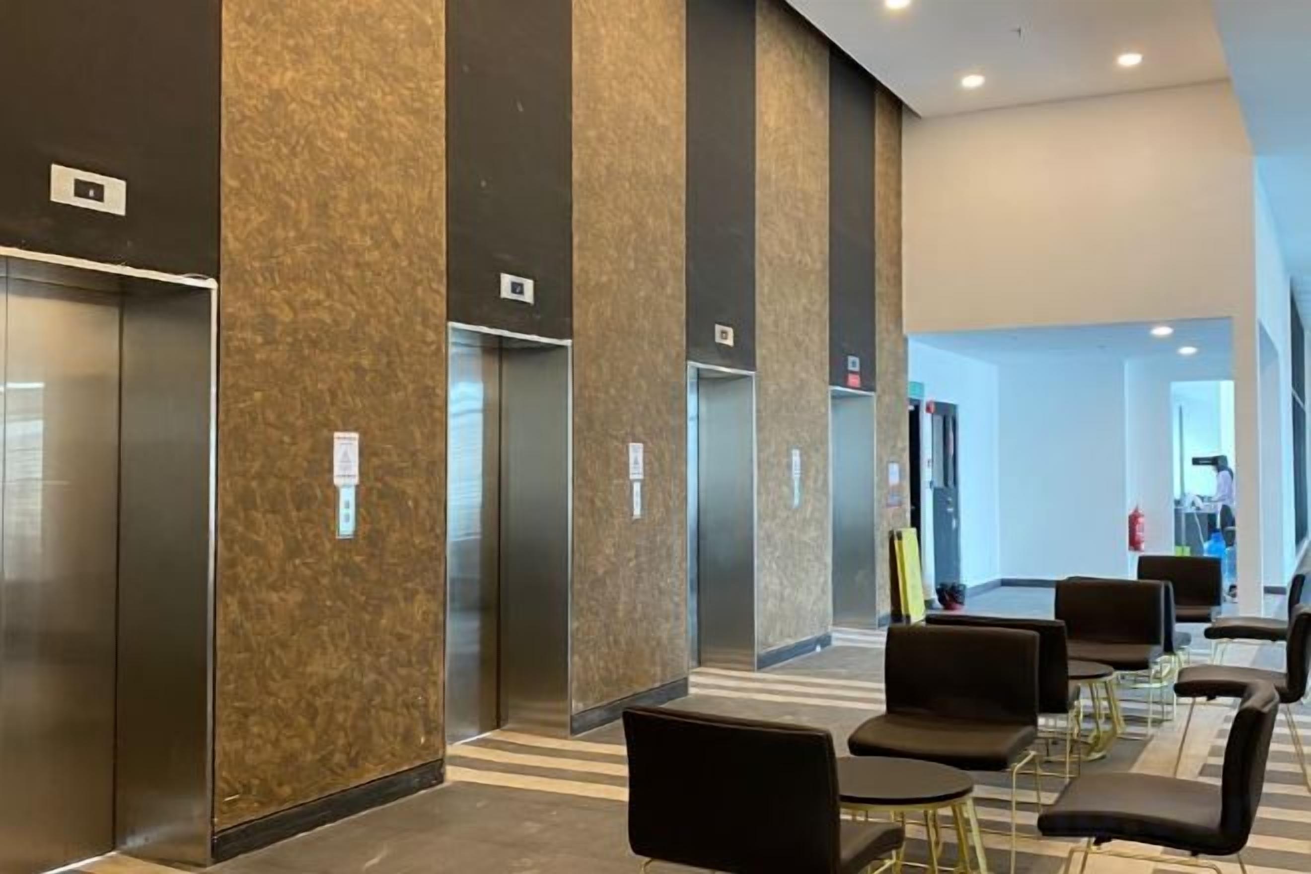 A modern elevator area with three elevators, a seating area with black chairs and tables, and a visible exit door.