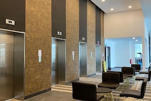 A modern elevator area with three elevators, a seating area with black chairs and tables, and a visible exit door. - Urban Suites Jelutong 3BR Apartment with Infinity Pool · Near George Town · 18min to Airport (George Town)