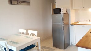 Een kleine keuken met een koelkast, magnetron en een houten aanrecht. Er is een eettafel met stoelen en een klein schilderij aan de muur.