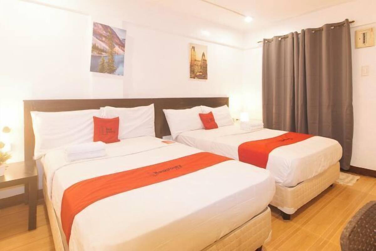 Een hotelkamer met twee bedden, elk voorzien van witte beddengoed en rode accenten, een houten hoofdbord en een groot raam met gordijnen.