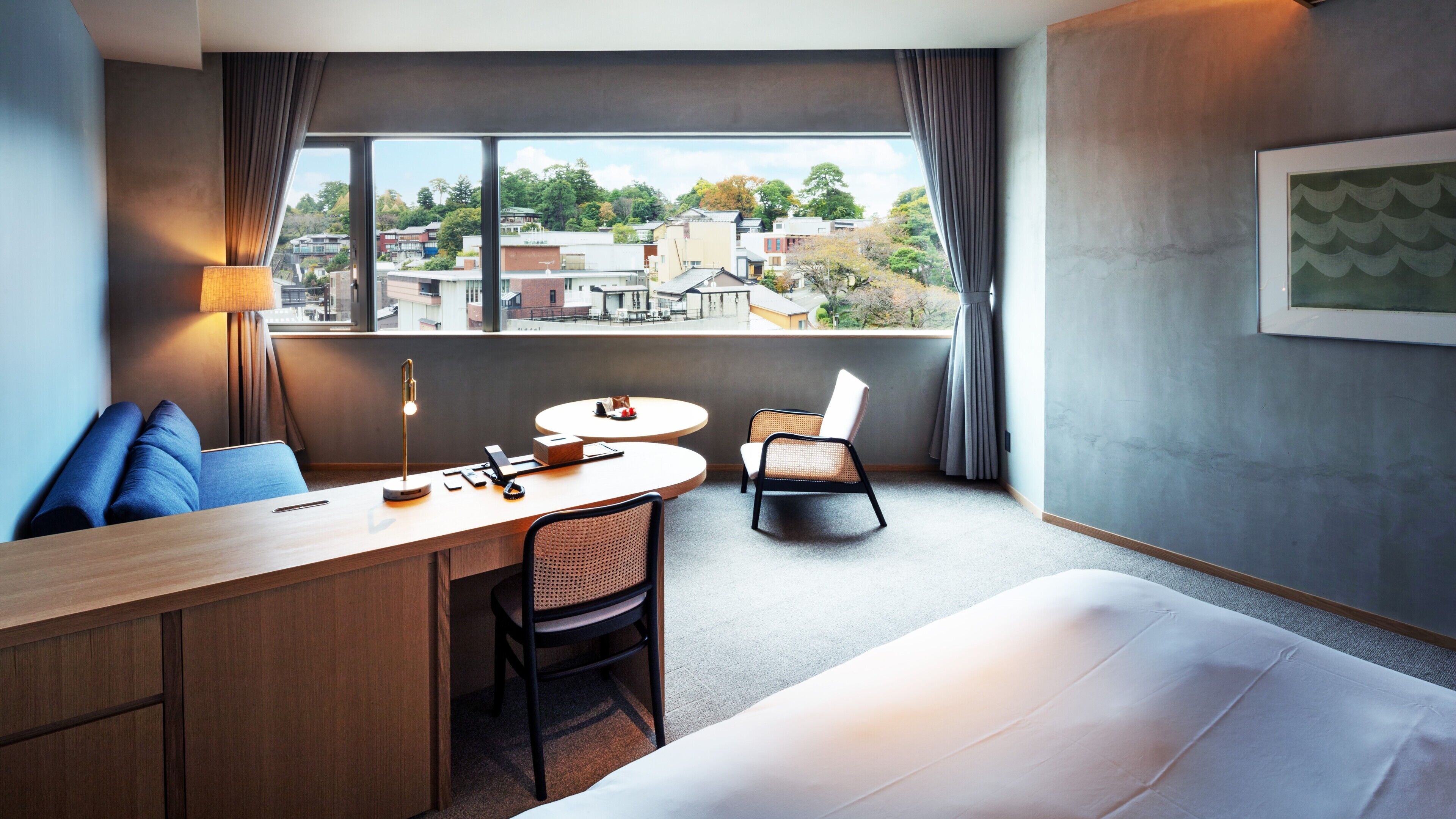 A hotel room with a bed, a desk, a chair, a table, and a view of the cityscape through large windows.