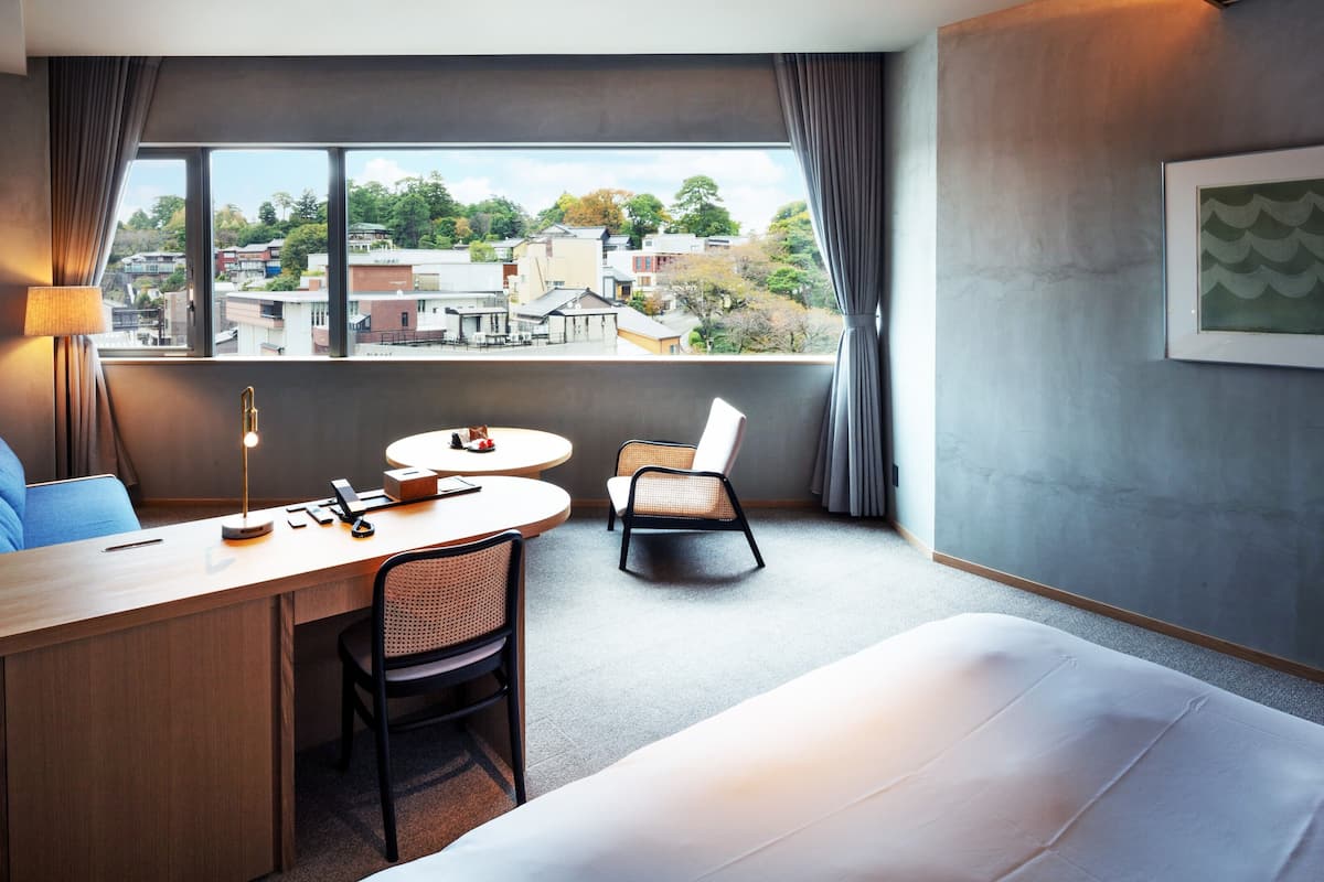 Une chambre d’hôtel équipée d’un lit, d’un bureau, d’une chaise, d’une table et offrant une vue sur le paysage urbain grâce à de grandes fenêtres.