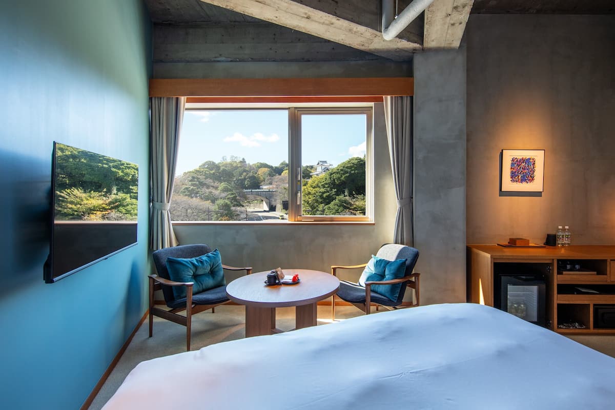 Une chambre d’hôtel avec un lit, deux chaises, une petite table, un bureau avec une lampe, un tableau au mur et une fenêtre donnant sur des arbres et un pont.