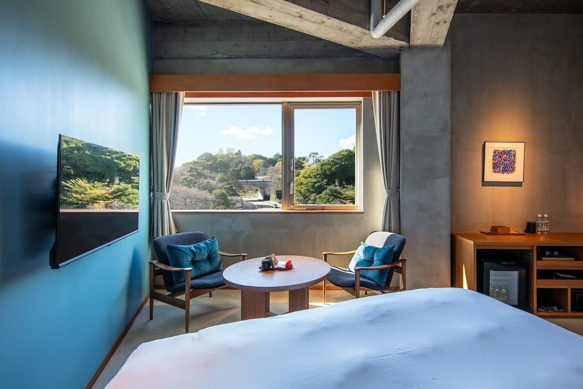 Une chambre d’hôtel avec un lit, deux chaises, une petite table, un bureau avec une lampe et une vue sur la verdure par la fenêtre.