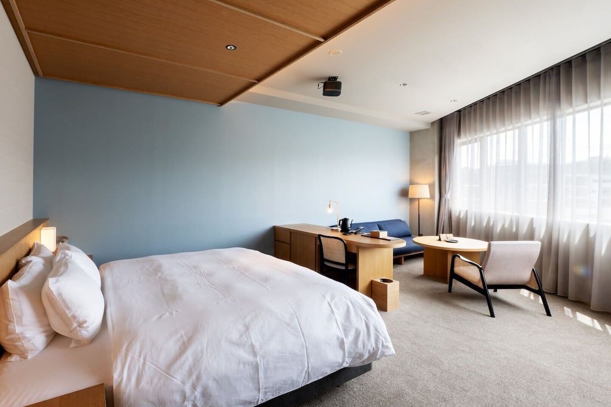 Une chambre d’hôtel avec un lit, un bureau, une chaise, une lampe et une grande fenêtre avec des rideaux.
