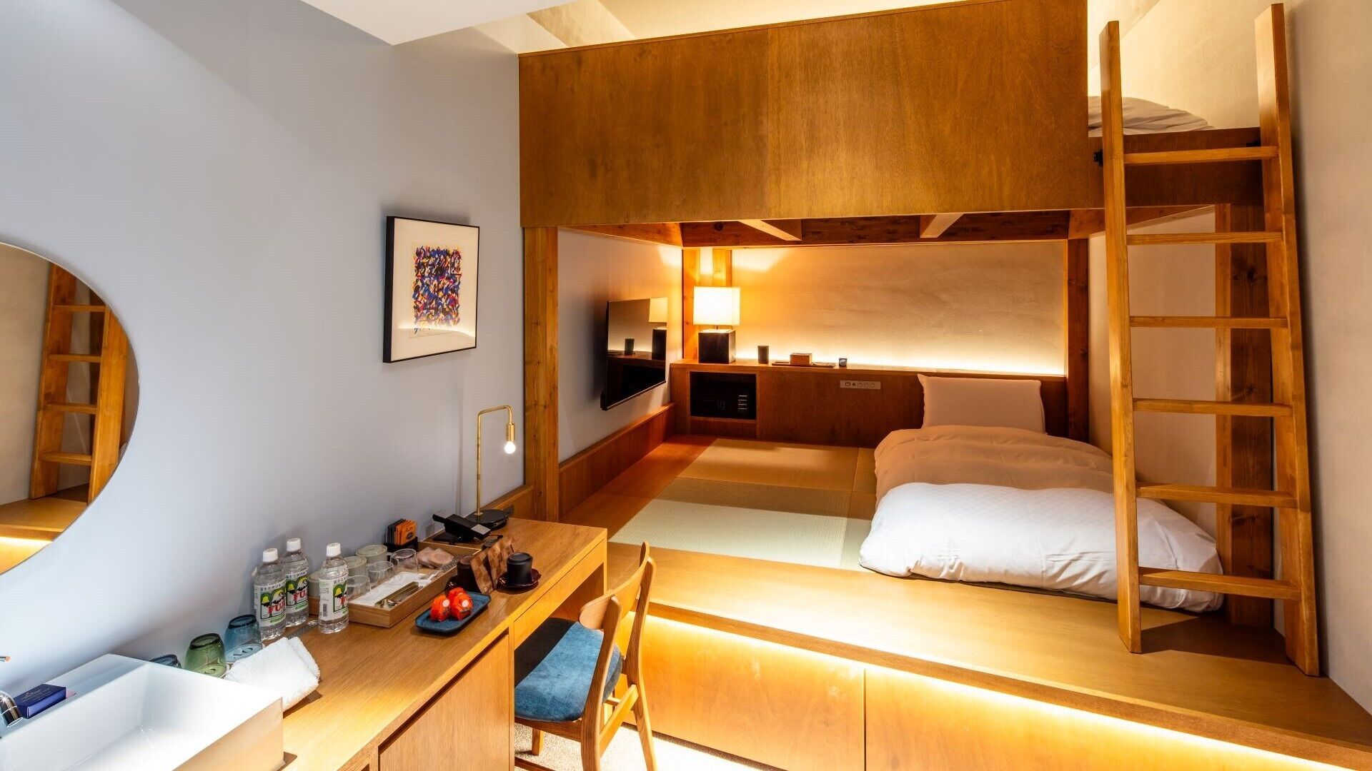 A compact hotel room with bunk beds, a desk with a chair, a small bathroom, and a bedside table with a lamp.