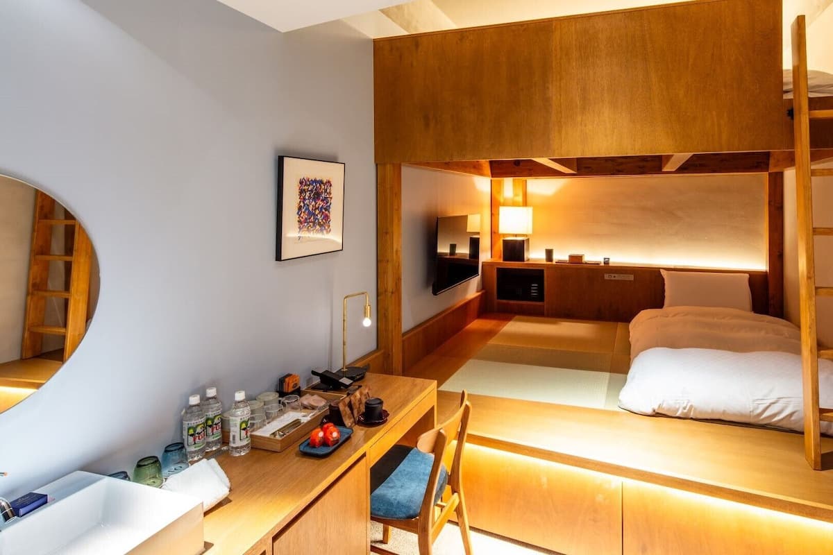 Une chambre d’hôtel compacte avec des lits superposés, un bureau avec une chaise, une petite salle de bain et une table de chevet avec une lampe.