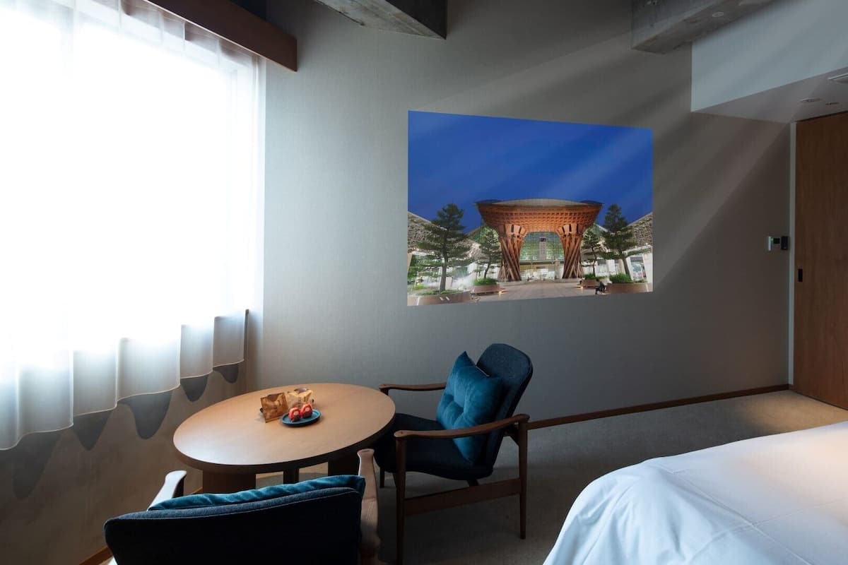 Une chambre d’hôtel avec un lit, une table ronde et une chaise, une fenêtre avec des rideaux et un tableau accroché au mur représentant un bâtiment.