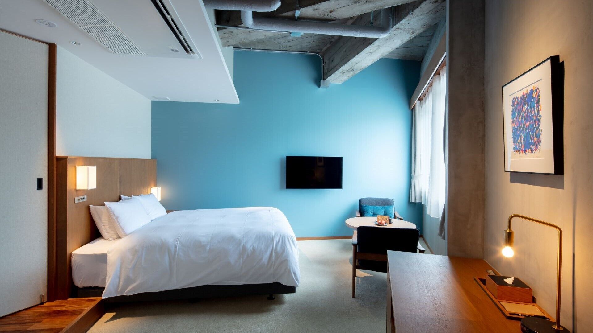 A hotel room with a bed, bedside table, chair, desk, TV, artwork, and exposed ceiling beams.
