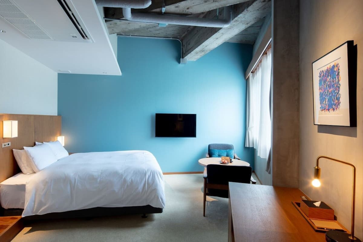 Une chambre d’hôtel comprenant un lit, une table de chevet, une chaise, un bureau, une télévision, des œuvres d’art et des poutres apparentes au plafond.