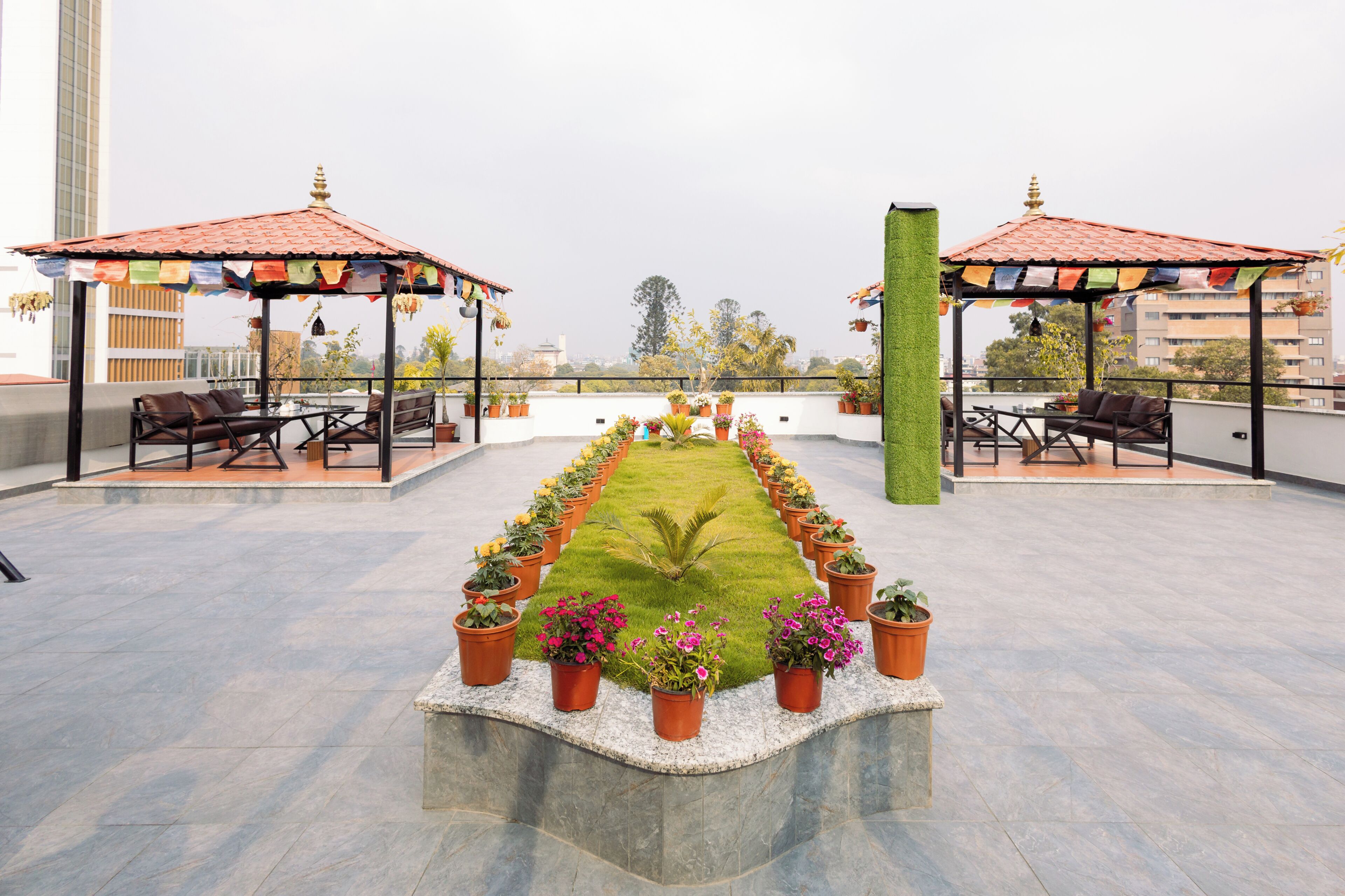 A rooftop terrace with a central lawn, two covered seating areas, and potted plants arranged in a line.