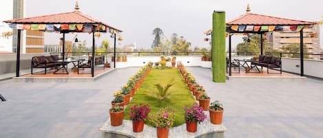 A rooftop terrace with a central lawn, two covered seating areas, and potted plants arranged in a line.