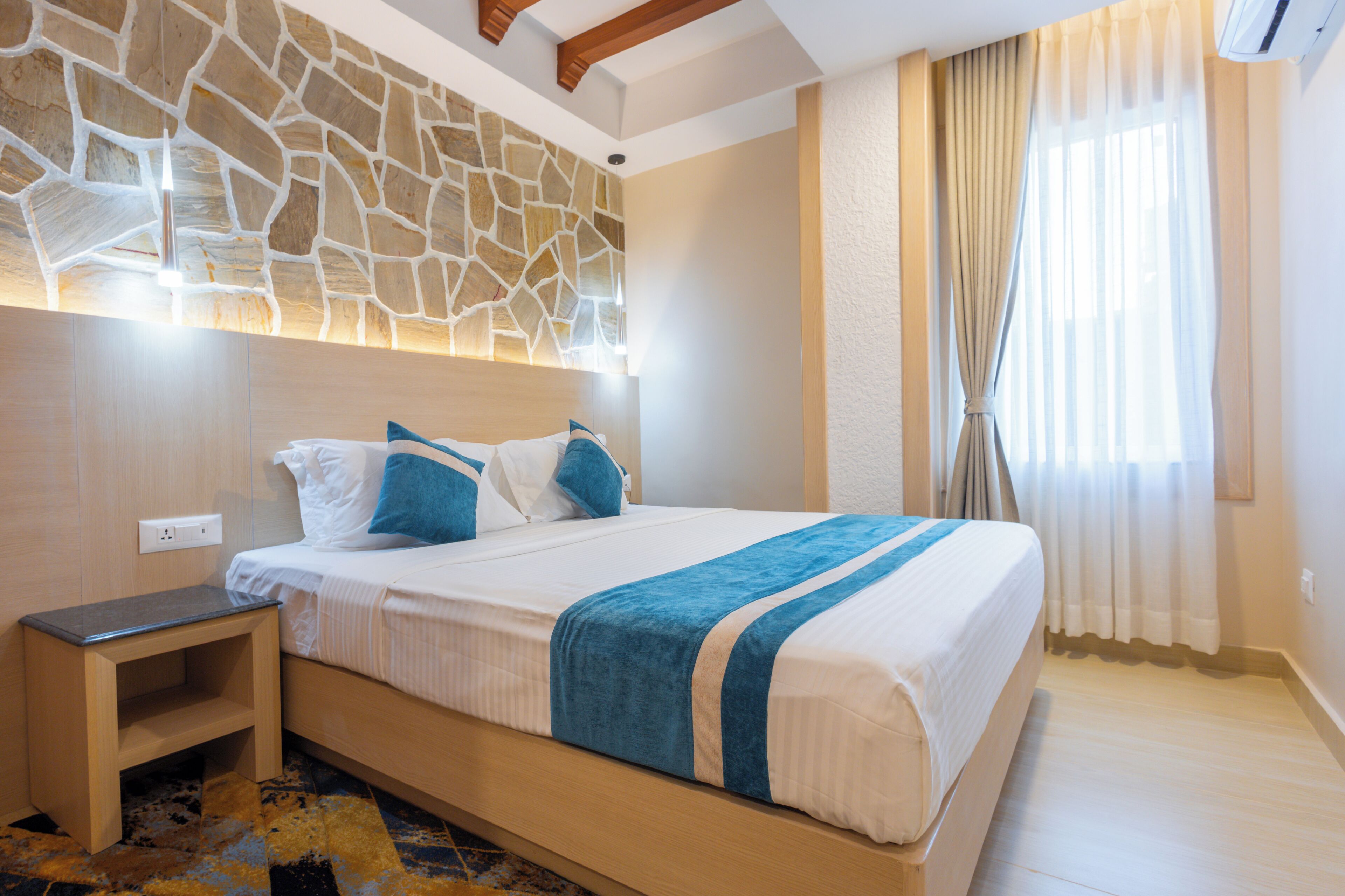 A bedroom with a stone wall, a bed with blue and white bedding, a nightstand, and a window with curtains.