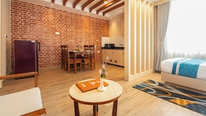A hotel room with a brick wall, wooden ceiling beams, a dining area, a kitchen, and a bed with a blue and white bedspread. - Tulsi Kathmandu Hotel (Kathmandu)
