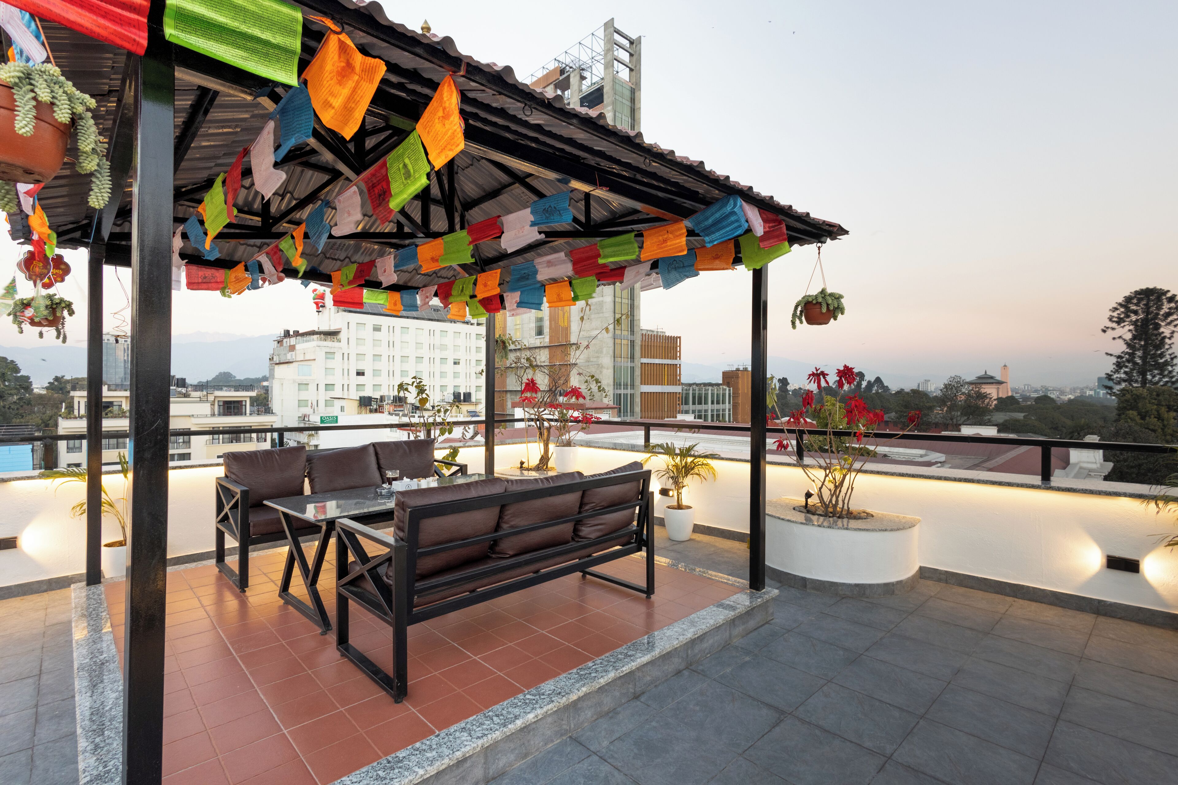 A rooftop terrace with a wooden bench and table, surrounded by colorful prayer flags, potted plants, and a view of the cityscape.