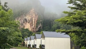 A row of simple, white, metal buildings with flat roofs, set against a backdrop of a misty cliff and lush greenery. - INK-PHA GARDENVIEW RESORT (Noen Maprang)