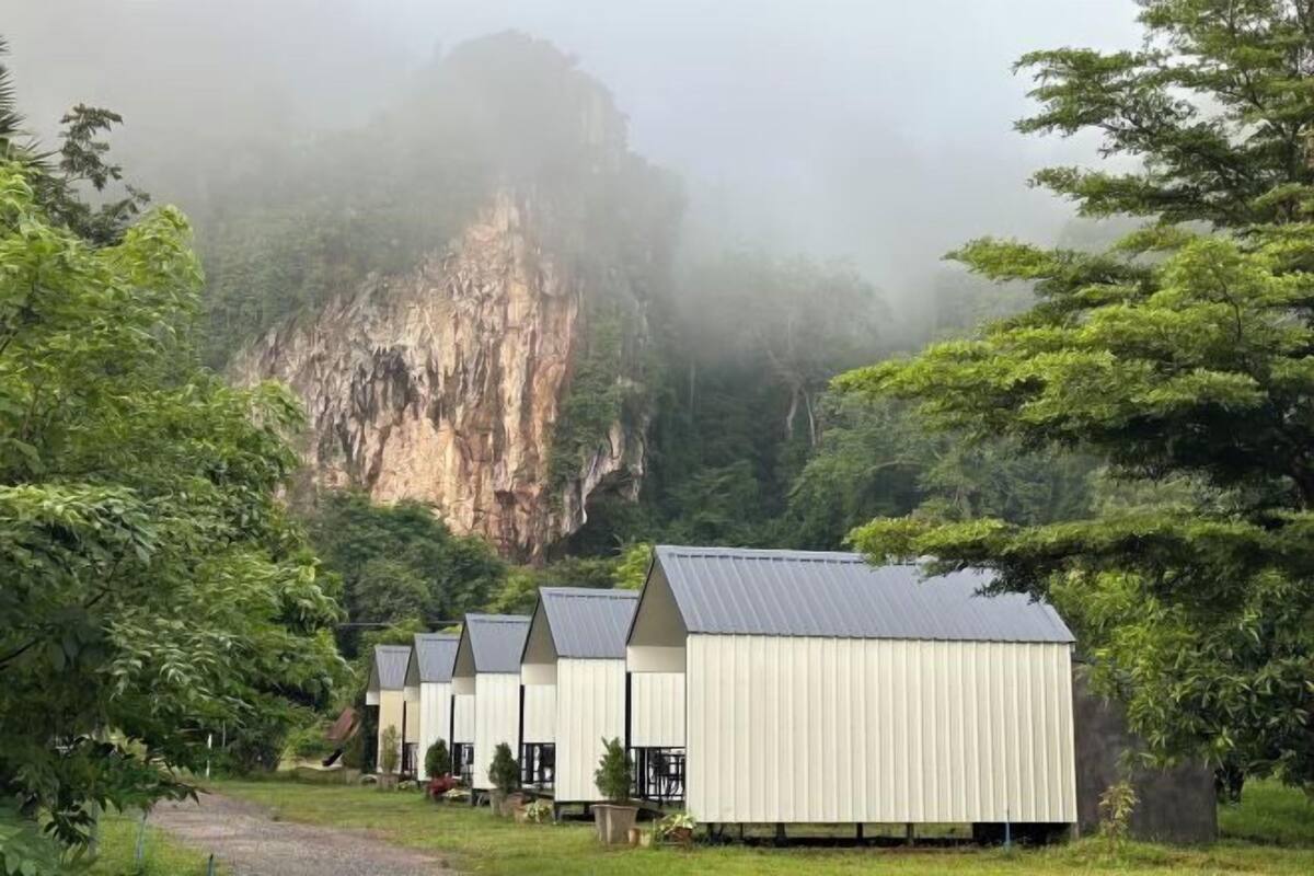A row of simple, white, metal buildings with flat roofs, set against a backdrop of a misty cliff and lush greenery.