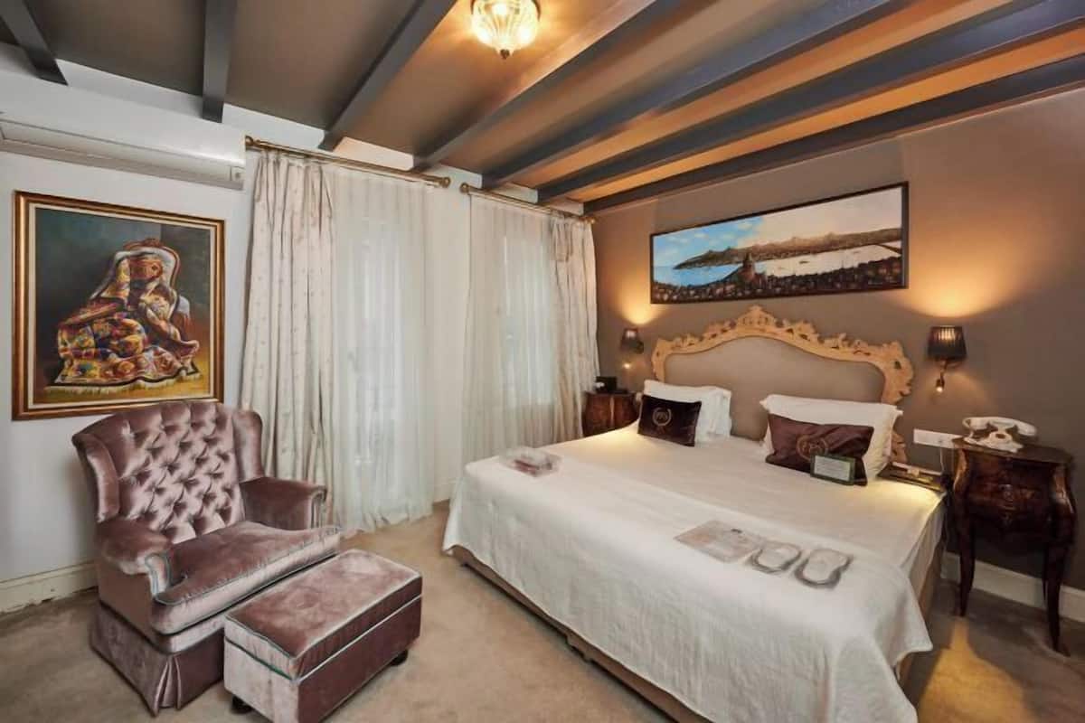 A bedroom with a large bed, a tufted armchair, a painting, and a view of a landscape.