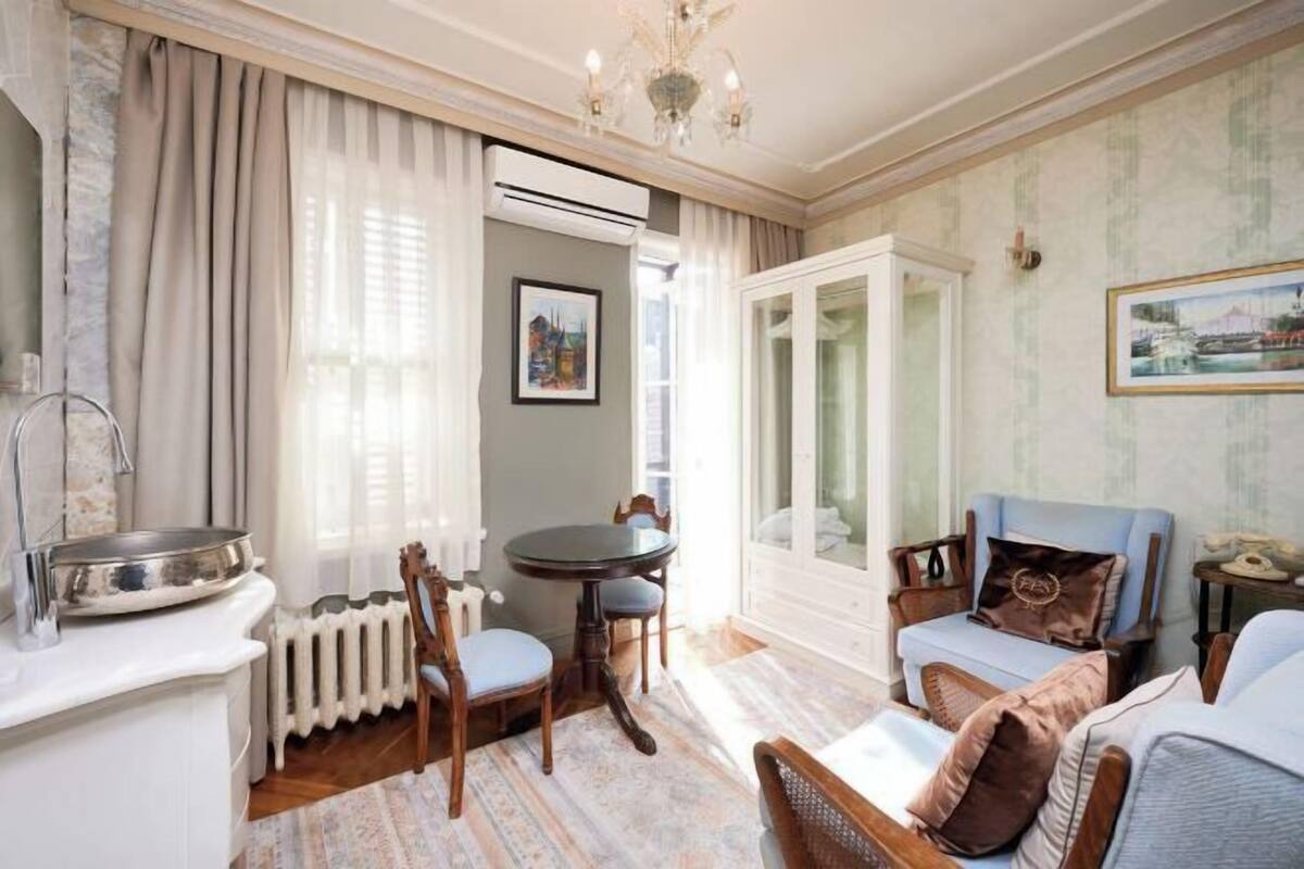 A room with a dining table, chairs, a sofa, a radiator, and a window with curtains.