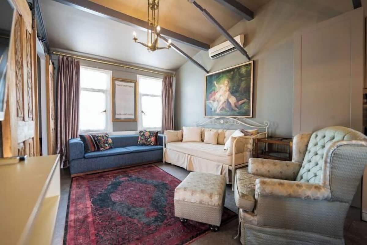 A cozy living room with a sofa, ottoman, and armchair, a large window with curtains, a painting on the wall, and a chandelier.