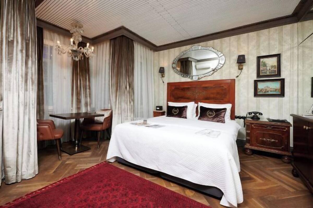 A hotel room with a bed, a desk, a chair, a red rug, a wooden headboard, a mirror, and framed pictures on the wall.