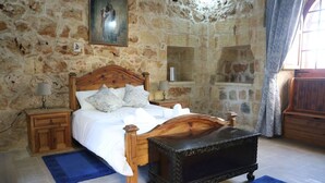 Une chambre avec un mur en pierre, un lit en bois avec une tête de lit, une table de chevet, une commode, un banc et une fenêtre avec des rideaux.