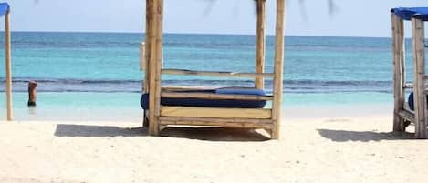 A beach with two wooden beach chairs with blue cushions, a thatched roof overhead, and a clear view of the ocean.