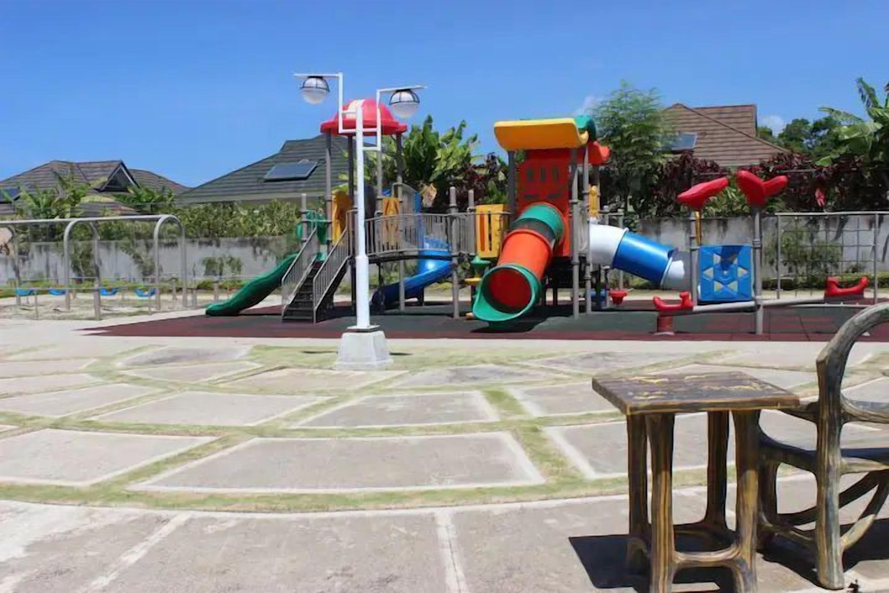 A playground with various play structures, including slides and climbing areas, surrounded by a paved area with benches.