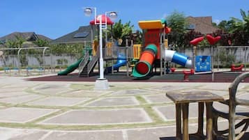 A playground with various play structures, including slides and climbing areas, surrounded by a paved area with benches.