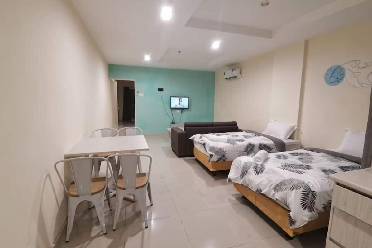 A room with a dining table, two beds with patterned bedding, a TV, and a wall-mounted air conditioner.