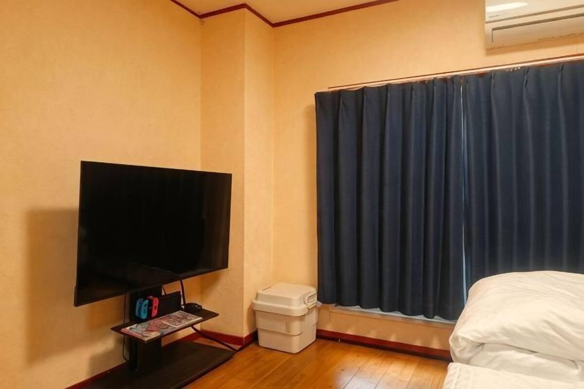 A room with a flat-screen TV, a small table with magazines, a window with dark curtains, and a bed.