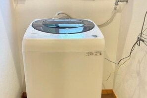 A compact, white washing machine with a transparent top, situated in a corner of a room with a plain wall and visible electrical wiring. - YiyashiHikari Fujiyoshida No.2 (Fujiyoshida)