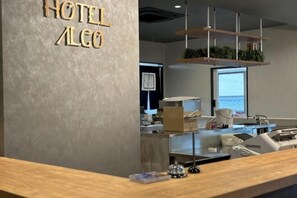 Un comptoir de réception d'hôtel avec une enseigne indiquant « HÔTEL ALGO », un ordinateur et une vue sur la mer par une grande fenêtre.