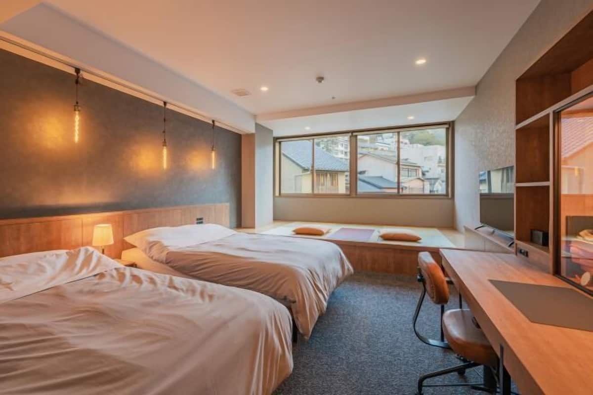 A hotel room with two beds, a desk, a chair, a large window, and a view of the outside.