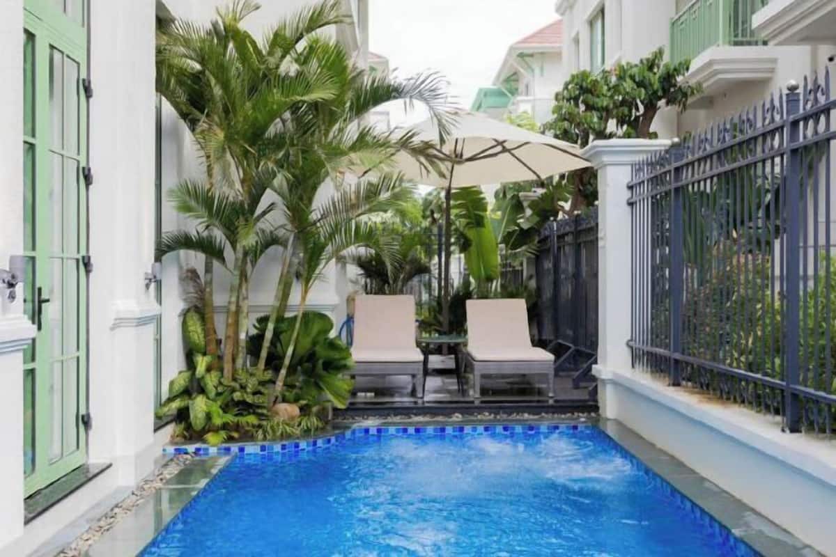 Un espace piscine bordé d’une mosaïque bleue, avec deux chaises longues et une table placée sous un parasol blanc.