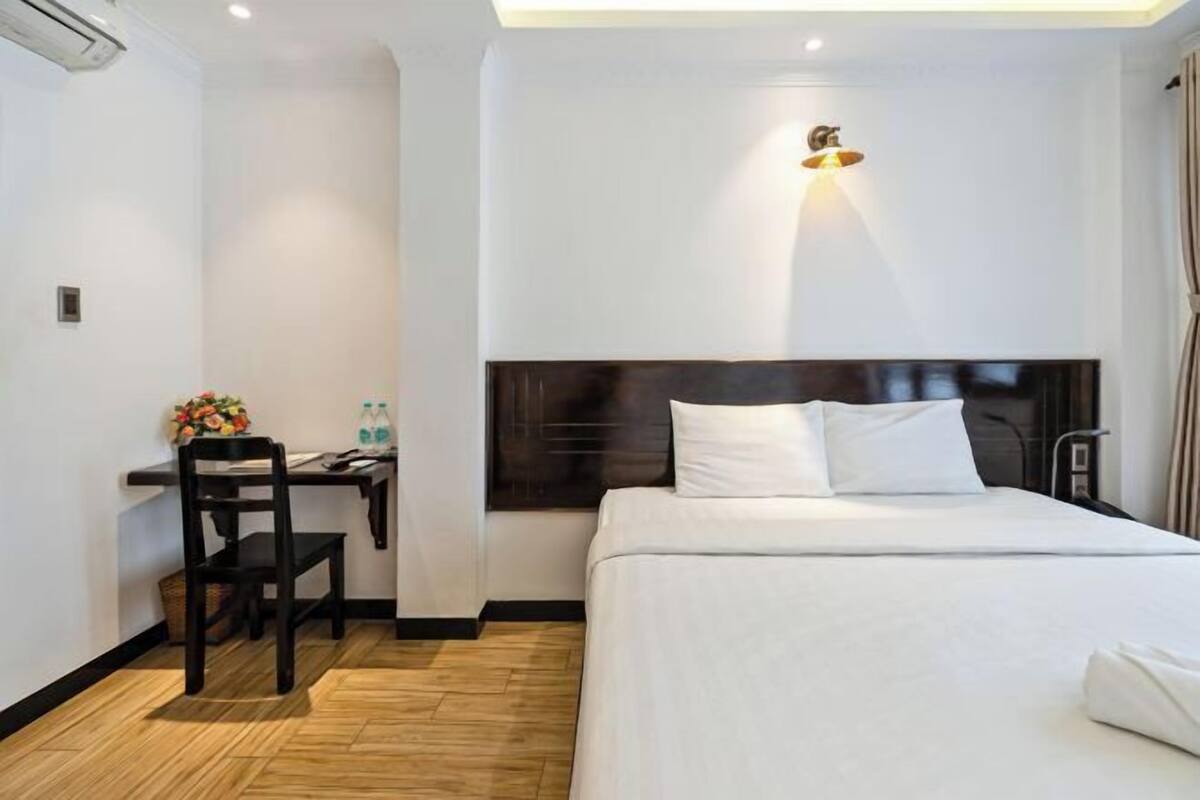Habitación de hotel con cama, escritorio con silla, una mesita con flores y una lámpara fijada a la pared.