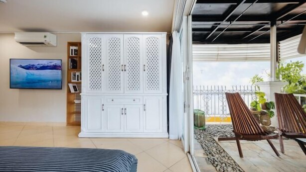A bedroom with a bed, a white wardrobe, a TV, and a balcony with chairs and plants. - VietHOME - 22 Pho Hue - Apartment 702 (Hanoi)