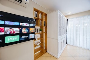 A modern interior with a flat-screen TV displaying various streaming services, a wooden bookshelf, and a large window with sheer curtains. - VietHOME - 22 Pho Hue - Apartment 702 (Hanoi)