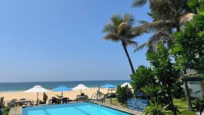 Una piscina con sillas de descanso y sombrillas, una playa al fondo y palmeras.
