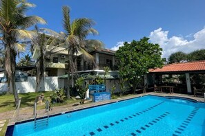 A swimming pool with clear blue water, surrounded by palm trees and a building with a red roof. - Pearl Island Beach Hotel (Hikkaduwa)