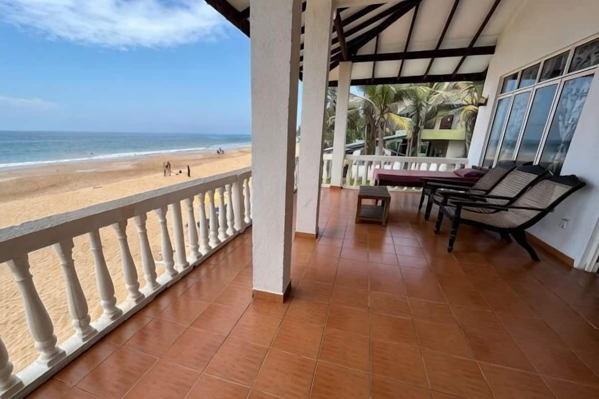 Een overdekt balkon met uitzicht op het strand, een zithoek en een hekwerk.