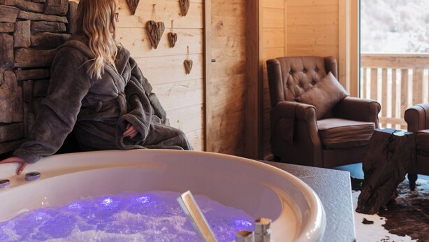 A wooden cabin interior with a stone wall, a large bathtub with purple lighting, and a brown leather chair.