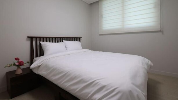 A bedroom with a bed, a nightstand, a vase with flowers, and a window with blinds. - EunhasuD&M Residence Hotel (Daejeon)