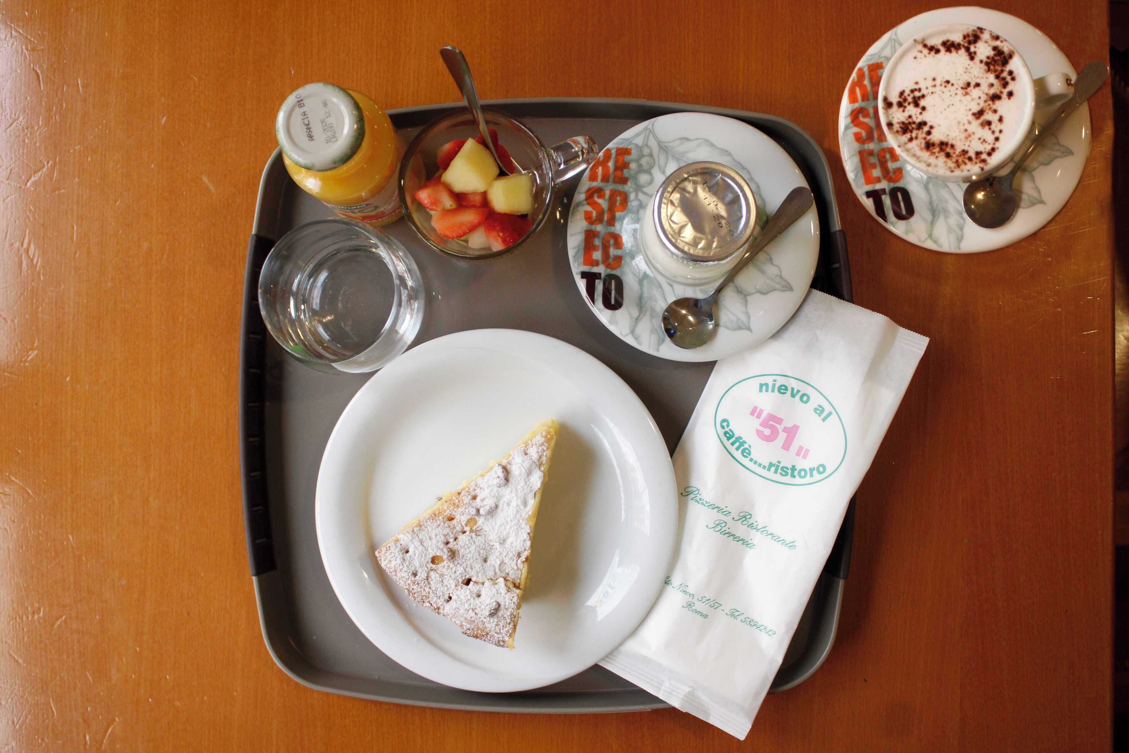 A tray with a slice of cake, a glass of water, a cup of coffee, a small bowl of fruit, and a bottle of juice.
