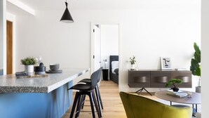 A modern kitchen with a granite countertop, black chairs, and a green armchair. There is a dining table with a potted plant and books. - Native Verso (London)
